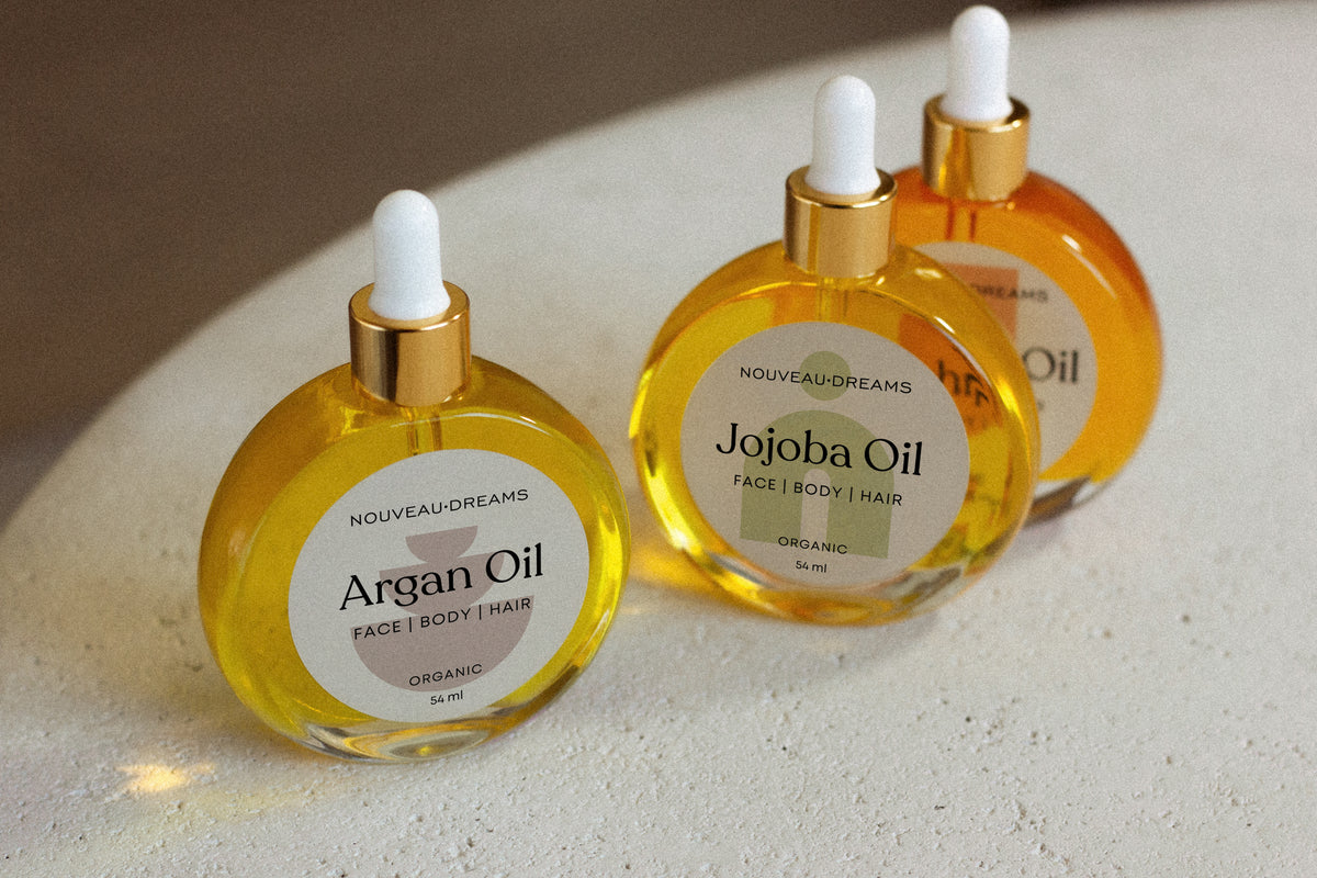 Close-up of Nouveau Dreams Argan Oil bottle in focus, with Jojoba and Rosehip Oils softly blurred in the background. All three are 54ml organic oils for face, body, and hair, in round glass dropper bottles.