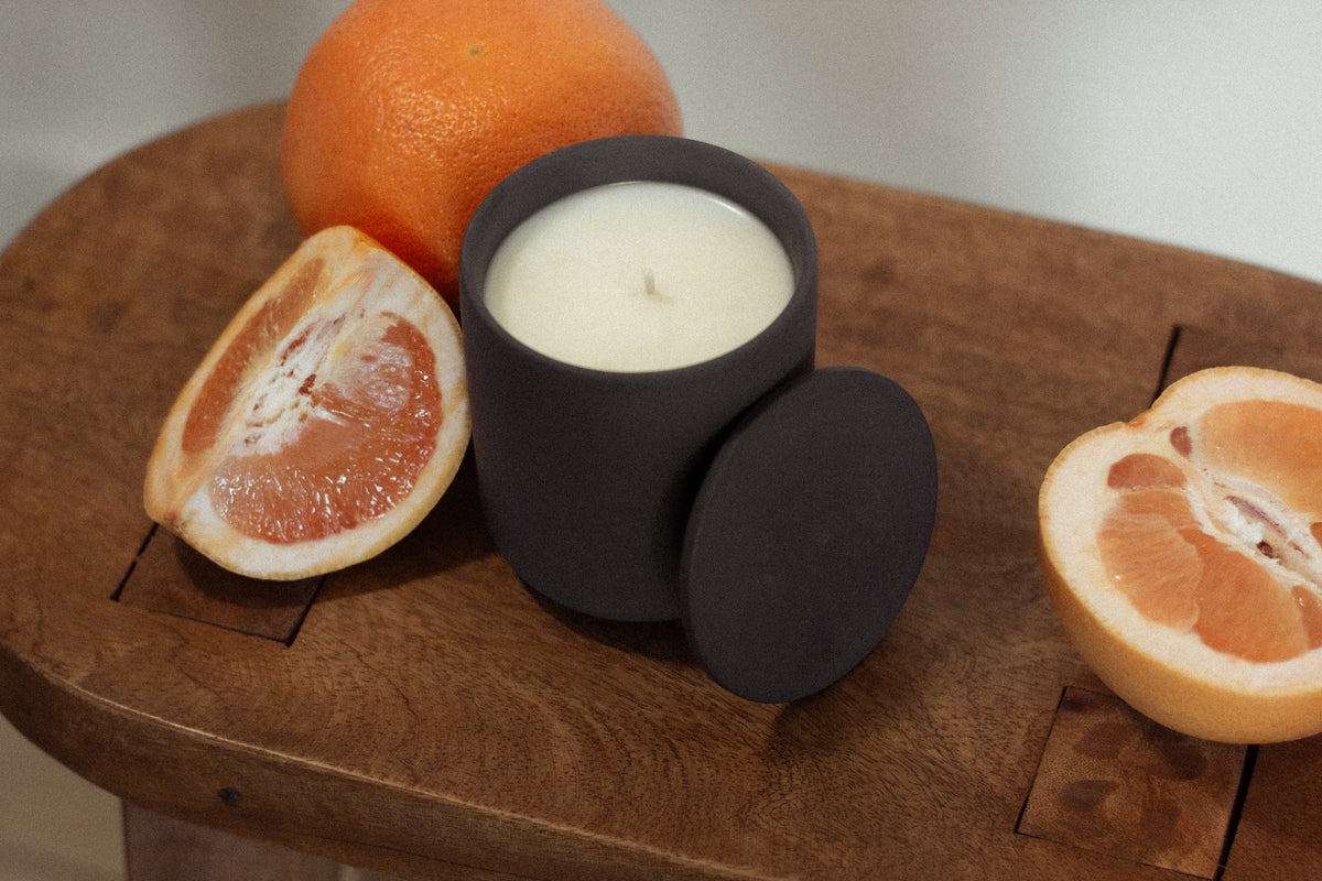 Unlit Grapefruit Vibrance candle in a matte black jar with lid, styled on a wooden surface beside fresh grapefruit halves