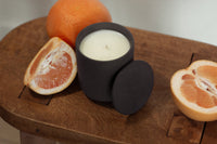 Unlit Grapefruit Vibrance candle in a matte black jar with lid, styled on a wooden surface beside fresh grapefruit halves