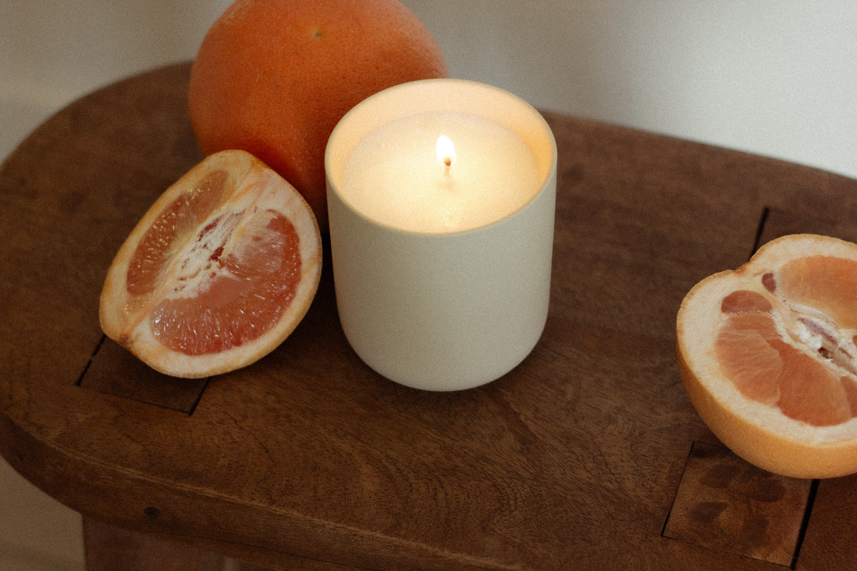 A lit Nouveau Dreams candle in a cream-colored vessel sits on a wooden surface, surrounded by fresh grapefruit halves and a whole grapefruit, evoking the scent of citrus and warmth