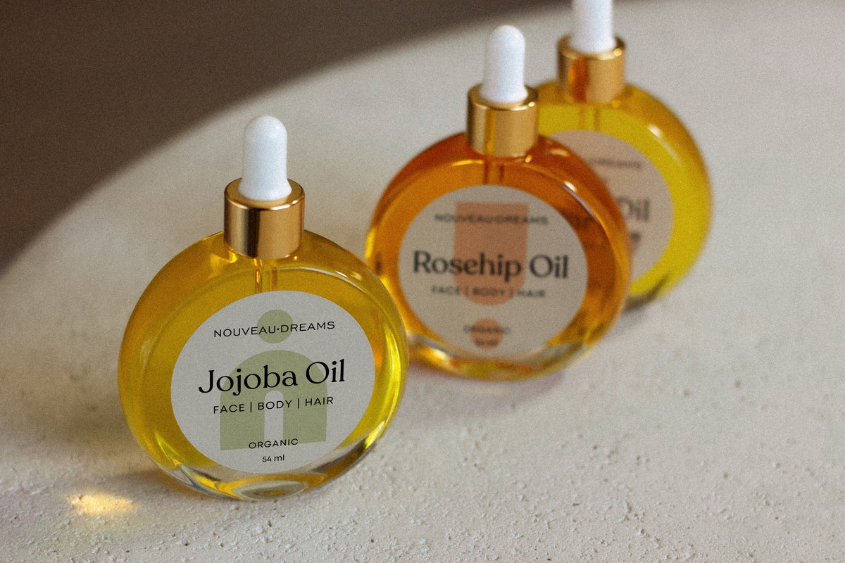 Front-facing Nouveau Dreams Jojoba Oil bottle with gold dropper in sharp focus, with Rosehip and Argan Oil bottles softly blurred in the background; all are 54ml organic oils for face, body, and hair.