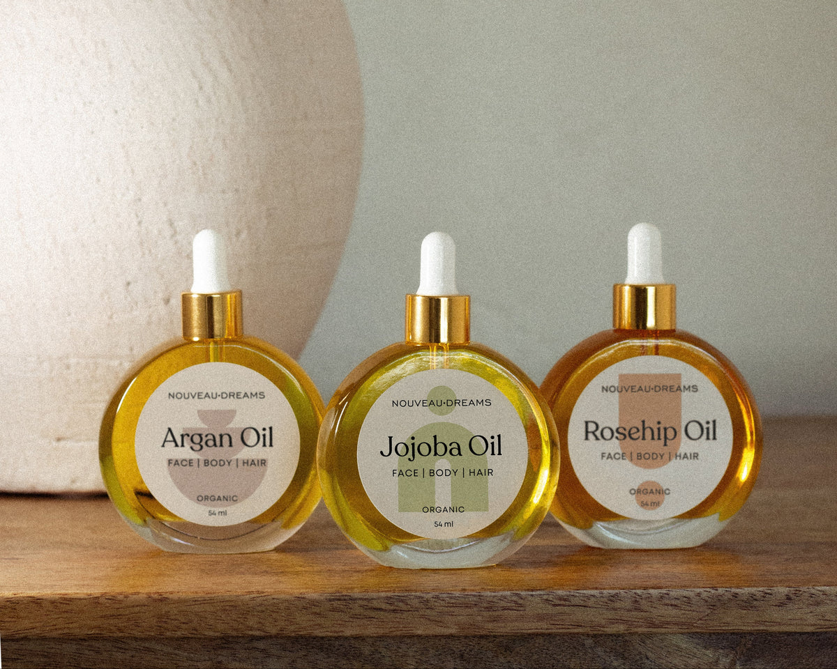 Three Nouveau Dreams organic oils—Argan, Jojoba, and Rosehip—in glass dropper bottles on a wooden surface beside a ceramic vase.
