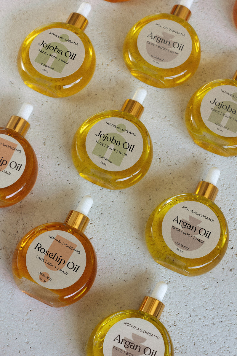 Flat lay of Nouveau Dreams organic oils in round glass bottles with dropper tops, including Argan, Jojoba, and Rosehip oil, 54ml each