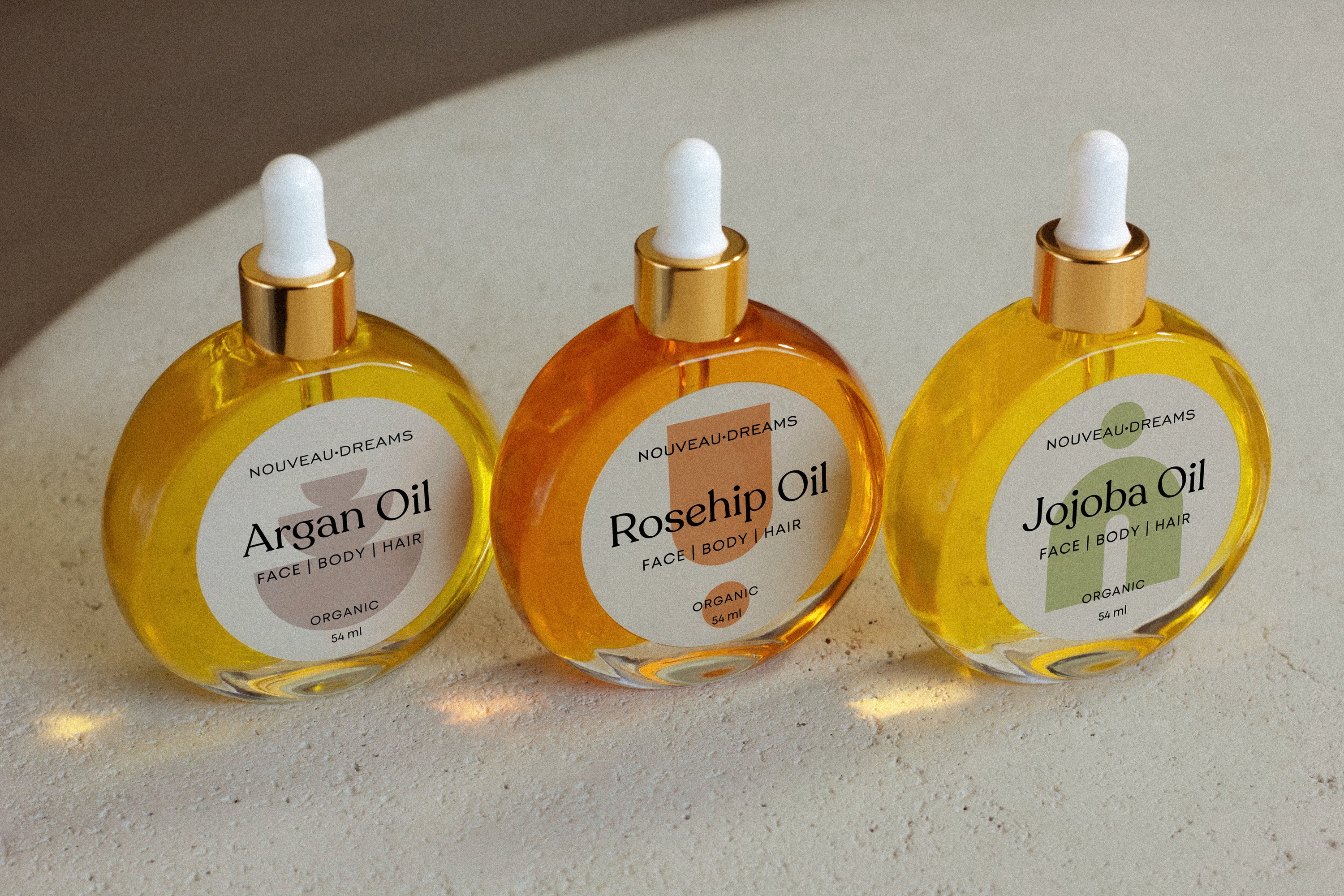 Three Nouveau Dreams organic carrier oils in round 54ml glass bottles with gold droppers — Argan Oil, Rosehip Oil, and Jojoba Oil — designed for face, body, and hair care.