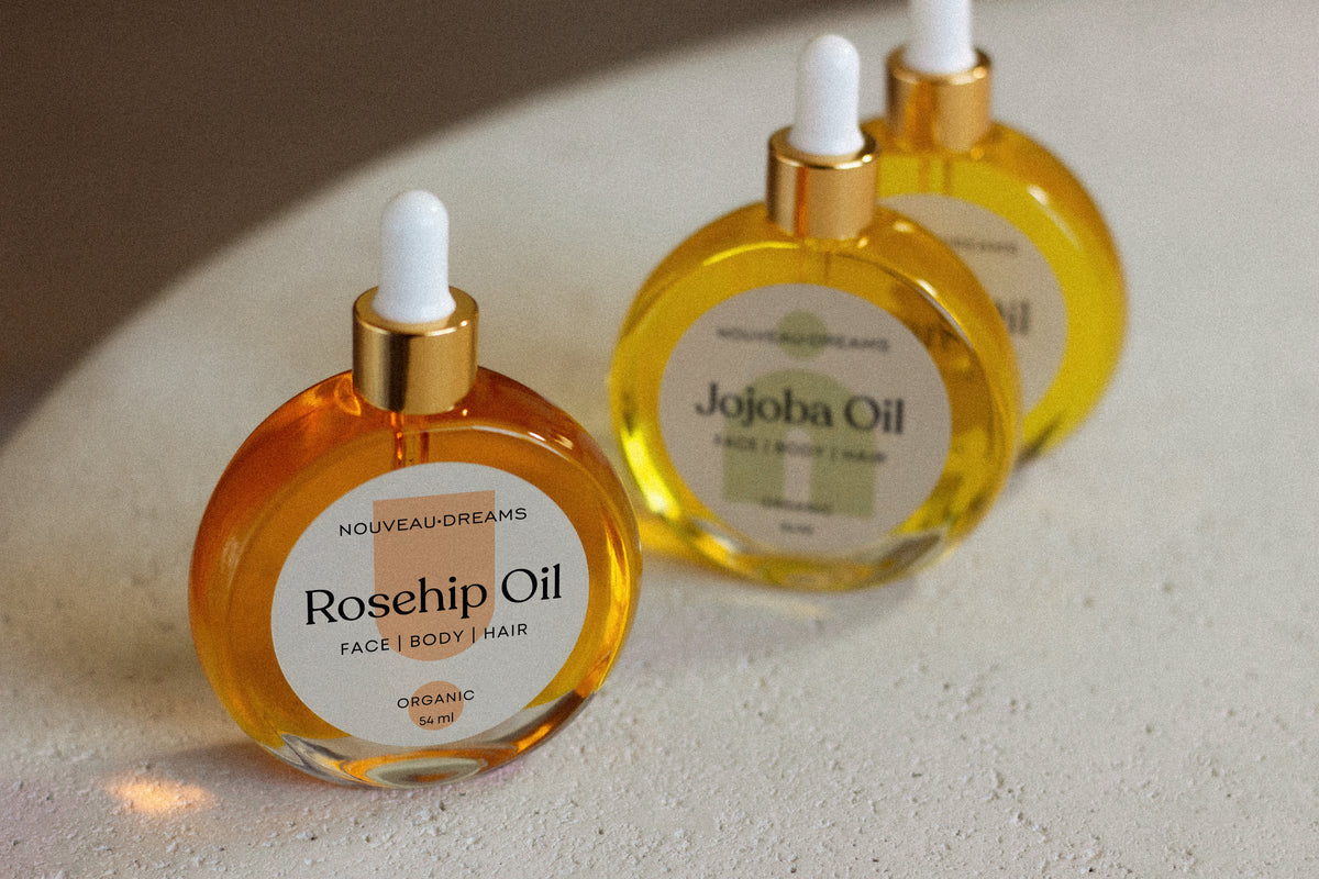 Close-up of Nouveau Dreams Rosehip Oil bottle in the foreground, with Jojoba and Argan Oils softly blurred in the background, all three are 54ml organic carrier oils for face, body, and hair.